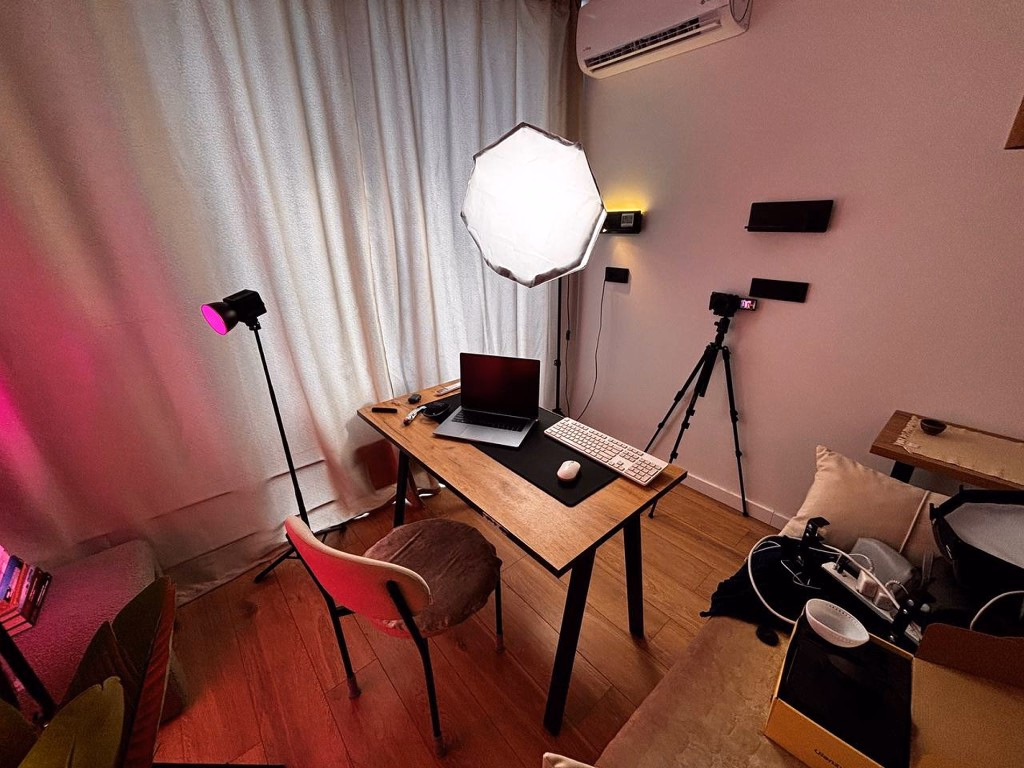 Setup caseiro de gravação: câmera em tripé, softbox e luz de apoio, notebook sobre a mesa, em um cômodo de casa